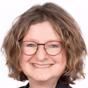 Porträt einer lächelnden Frau mit lockigem, braunem Haar und Brille vor einem weißen Hintergrund.