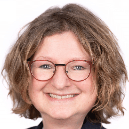 Porträt einer lächelnden Frau mit lockigem, braunem Haar und Brille vor einem weißen Hintergrund.