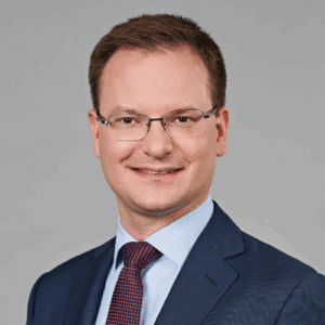 Porträt eines Mannes mit Brille, braunen Haaren, in einem Anzug mit Krawatte, vor einem neutralen Hintergrund.