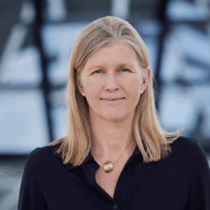 Porträt einer Frau mit blonden Haaren, die in einem schwarzen Oberteil lächelt, vor einem unscharfen Hintergrund.