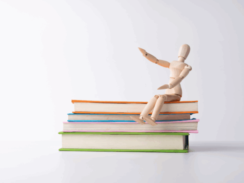 Holzfigur sitzt auf einem Stapel bunter Bücher und hebt eine Hand, vor neutralem Hintergrund.