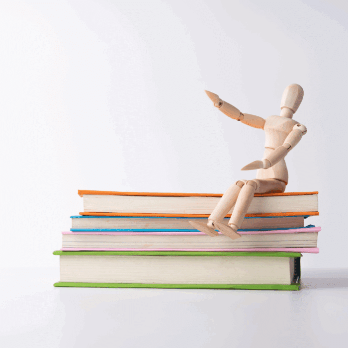 Holzfigur sitzt auf einem Stapel bunter Bücher und hebt eine Hand, vor neutralem Hintergrund.