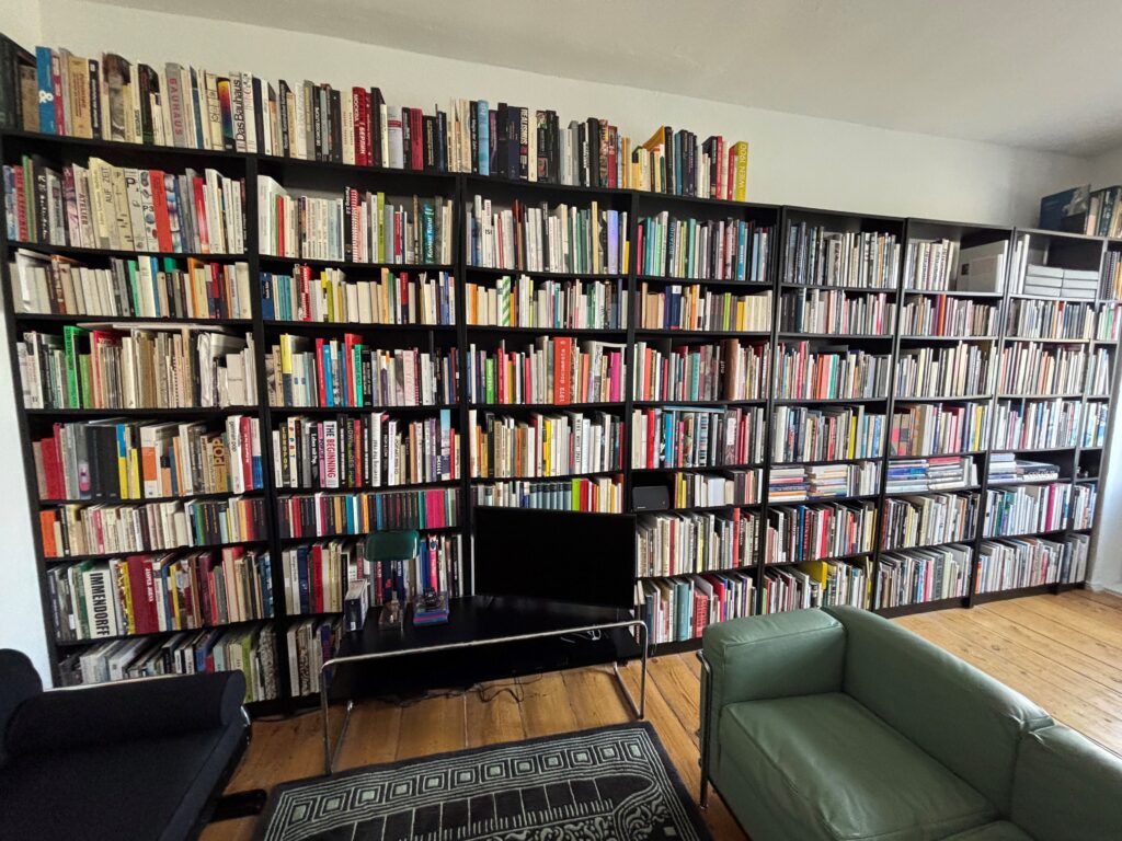 | Human Resources Manager Bücherwand mit zahlreichen bunten Buchrücken, schwarzem Regal und einem grünen Sessel im Vordergrund.