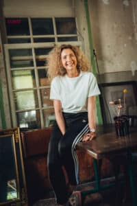 Lächelnde Frau mit lockigem Haar sitzt auf einem Tisch in einem rustikalen Raum, umgeben von Spiegeln und Blumen.
