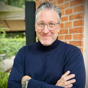 Mann mit Brille und dunklem Rollkragenpullover, lächelt und steht mit verschränkten Armen vor einer Backsteinwand.