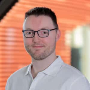 Porträt eines Mannes mit Brille, kurzen Haaren und einem weißen Polohemd, vor einem orangefarbenen Hintergrund.