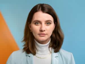 Porträt einer Frau mit braunen Haaren, blauer Bluse und hellem Rollkragenpullover, vor einem farbigen Hintergrund.