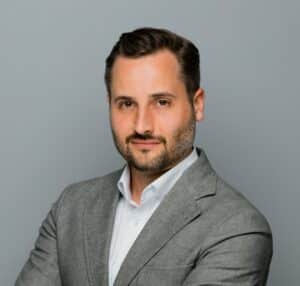 Mann mit kurzem Haar und Bart, trägt einen grauen Anzug und schaut direkt in die Kamera vor einem grauen Hintergrund.