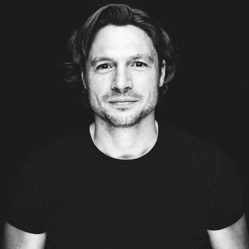 Porträt eines Mannes mit kurzen, lockigen Haaren, der in die Kamera lächelt, vor einem schwarzen Hintergrund.