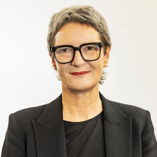 Porträt einer Frau mit kurzen grauen Haaren, Brille und schwarzem Blazer, lächelnd vor neutralem Hintergrund.