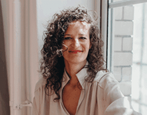 Frau mit lockigem Haar und rotem Lippenstift, lächelt vor einem Fenster in einem hellen Raum.