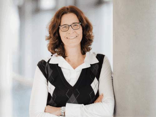 Frau mit lockigem Haar und Brille, lächelt, steht an einer Säule, trägt einen schwarzen Pullover mit Rautenmuster und e...