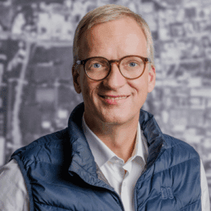 Mann mit Brille und blauer Weste lächelt vor einem unscharfen, hellen Hintergrund.