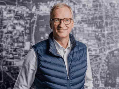 Mann mit Brille und blauer Weste lächelt vor einer verschwommenen, grafischen Hintergrundlandschaft.