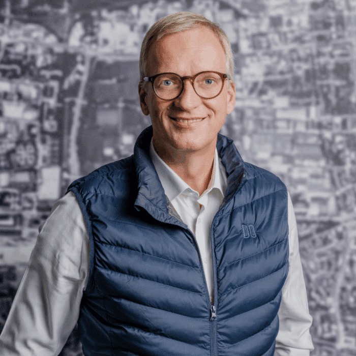 Mann mit Brille und blauer Weste lächelt vor einer verschwommenen, grafischen Hintergrundlandschaft.
