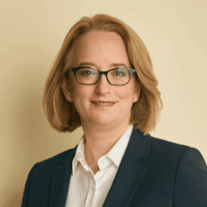 katharina-hermann Porträt einer Frau mit blonden Haaren, Brille und einem dunklen Blazer, vor einem hellen Hintergrund.