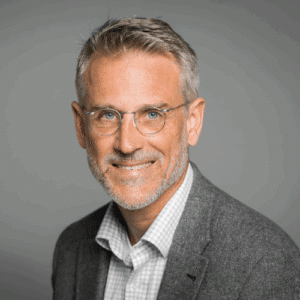 Porträt eines lächelnden Mannes mit Brille, grauem Haar und Bart, in einem grauen Anzug vor neutralem Hintergrund.