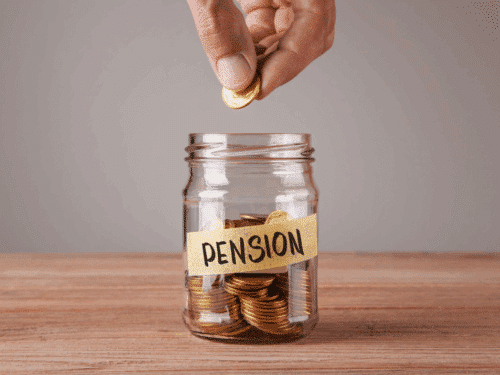 Hand legt Münze in ein Glas mit der Beschriftung „PENSION“, gefüllt mit weiteren Münzen auf einem Holztisch.