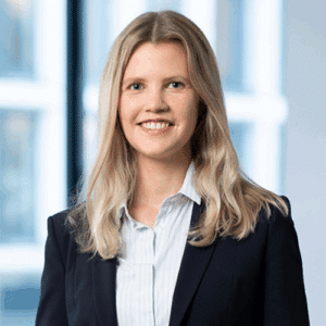 Porträt einer lächelnden Frau mit langen blonden Haaren, in einem schwarzen Blazer und blauen Hemd, vor einem hellen Hin...