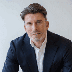 Mann mit kurzen, grauen Haaren und Bart, trägt einen dunklen Anzug und schaut direkt in die Kamera.