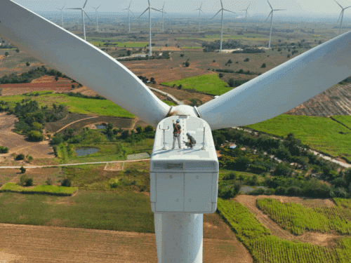 Beitragsbild Website Blick von oben auf einen Windkraftanlagenrotor mit Arbeitern, umgeben von grünen Feldern und weiteren Windrädern im Hint...