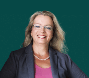 Frau mit langen, blonden Haaren, Brille und lächelndem Gesicht, trägt einen Blazer über einem pinken Oberteil.