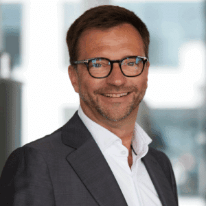 Personenkasten Lächelnder Mann mit Brille in Anzug, vor unscharfem Hintergrund.