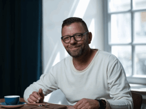 Mann mit Brille und kurzem Bart sitzt an einem Tisch, lächelt und hält eine Brille in der Hand, Tasse daneben.