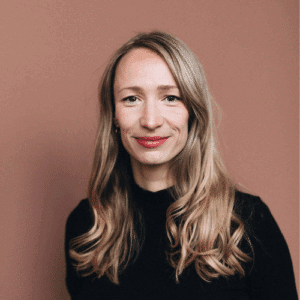 Porträt einer lächelnden Frau mit langen, blonden Haaren vor einem rosa Hintergrund. Sie trägt ein schwarzes Oberteil.