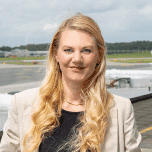 Frau mit langen blonden Haaren, trägt einen hellen Blazer, steht vor einem Flughafen mit Flugzeugen im Hintergrund.