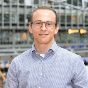 Junger Mann mit Brille und Hemd lächelt vor einer modernen, lichtdurchfluteten Umgebung.