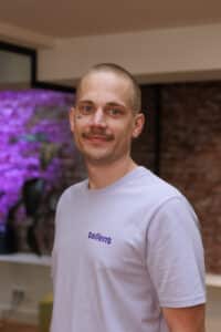 Mann mit kurzem Haar und Schnurrbart, trägt ein lila T-Shirt mit dem Aufdruck "tellent", vor einer Backsteinwand.