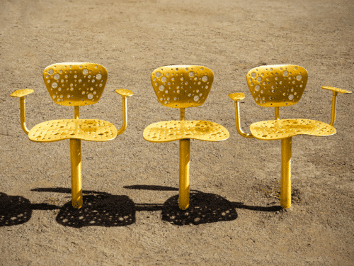 Drei gelbe Stühle mit Armlehnen stehen auf einem sandigen Boden, Schattenmuster fallen auf die Fläche.