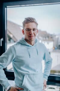 Junger Mann mit Brille und hellblauem Hoodie steht vor einem Fenster mit urbanem Hintergrund.