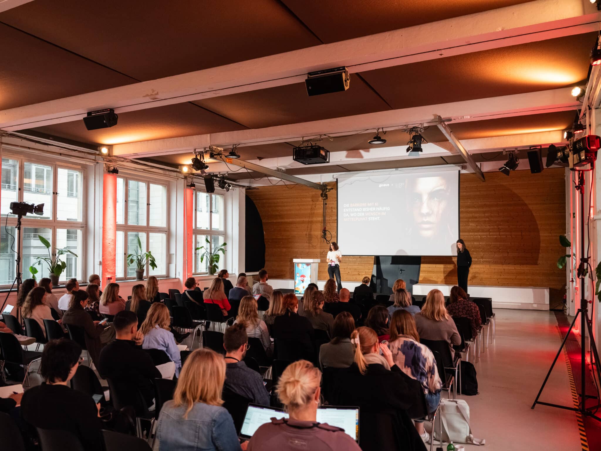 Konferenzraum mit Publikum, Vortragende auf der Bühne, Präsentation im Hintergrund, Pflanzen und modernes Ambiente.