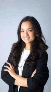 Junge Frau mit langen, lockigen Haaren in schwarzer Jacke, lächelt und steht mit verschränkten Armen vor grauem Hintergr...