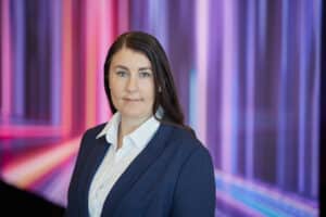 Frau mit langen, dunklen Haaren in einem blauen Blazer vor einem bunten, abstrakten Hintergrund.