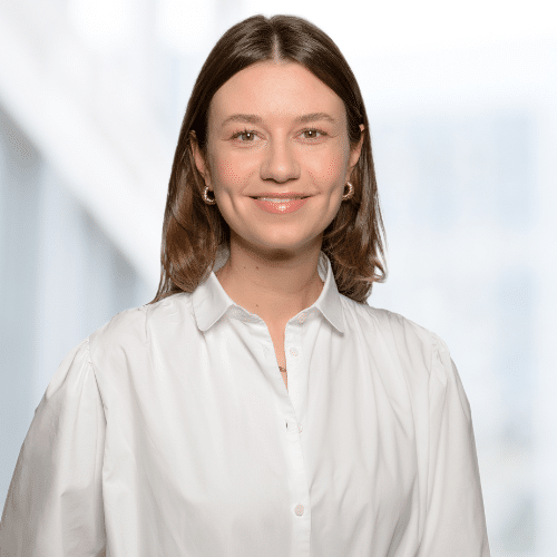 Porträt einer lächelnden Frau mit braunen Haaren, die ein weißes Hemd trägt, vor einem hellen, unscharfen Hintergrund.