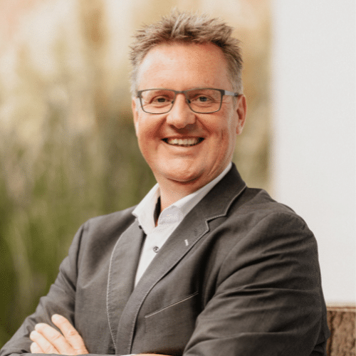 Lächelnder Mann in Brille, mit verschränkten Armen, trägt einen grauen Anzug vor unscharfem, grünem Hintergrund.