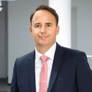 Porträt eines Mannes in Anzug mit hellblauer Krawatte, lächelnd vor einem modernen, hellen Hintergrund.