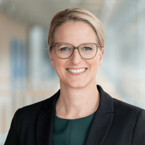 Porträt einer lächelnden Frau mit Brille, in einem schwarzen Blazer, vor einem hellen, modernen Hintergrund.