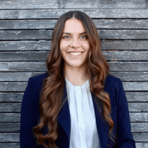 Frau mit langen, lockigen Haaren in einem blauen Blazer vor einer Holzlattenwand, lächelt freundlich in die Kamera.