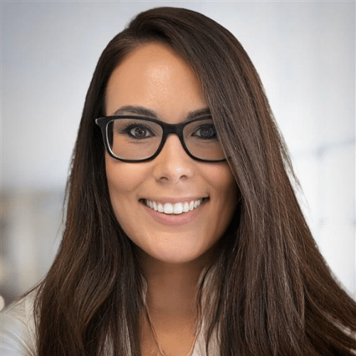 Lächelnde Frau mit langen, braunen Haaren und Brille, vor einem hellen, unscharfen Hintergrund.