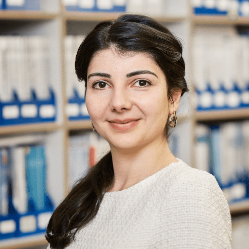 Porträt einer Frau mit dunklen Haaren, die in einem hellen Pullover vor einem Bücherregal lächelt.
