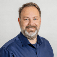 Porträt eines lächelnden Mannes mit Bart, in einem blauen Hemd, vor einem hellen Hintergrund.