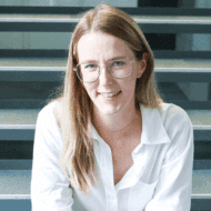 Junge Frau mit langen blonden Haaren und Brille, sitzt auf einer Treppe und lächelt in die Kamera.