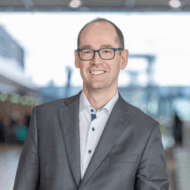 Mann in Anzug mit Brille lächelt vor einem modernen Flughafen-Hintergrund.