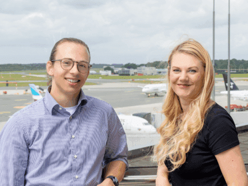 Zwei Personen lächeln vor einem Flughafen-Hintergrund mit Flugzeugen und Wolken am Himmel.