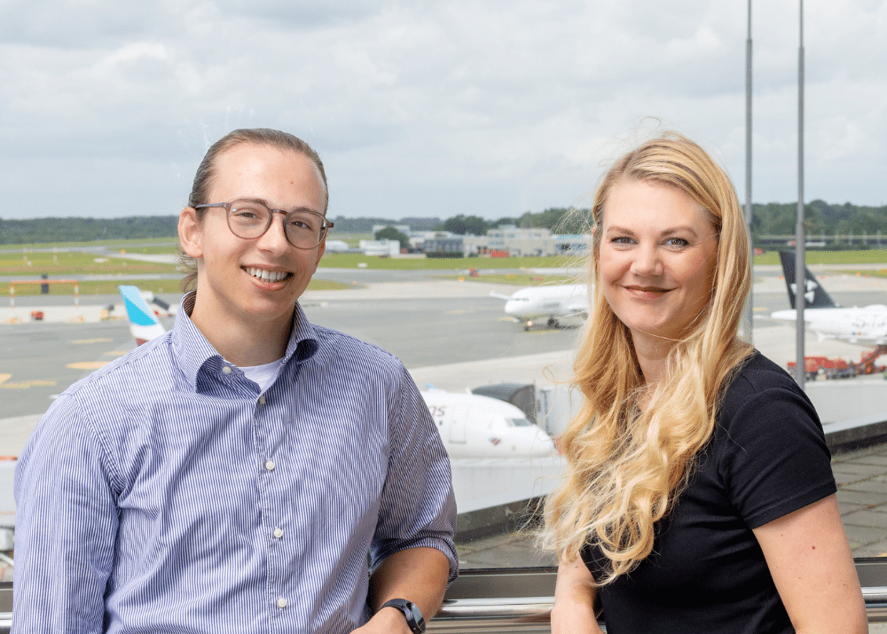 Zwei Personen lächeln vor einem Flughafen-Hintergrund mit Flugzeugen und Wolken am Himmel.