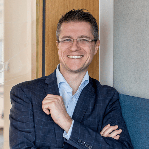 Mann mit Brille und Anzug lächelt, steht an einer Wand mit Holz- und Stoffelementen.